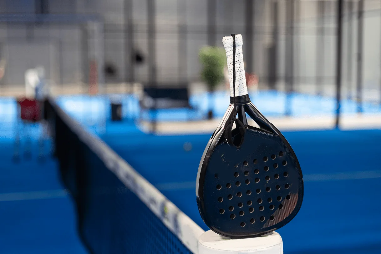 Black padel racket resting on a net post beside a blue indoor court