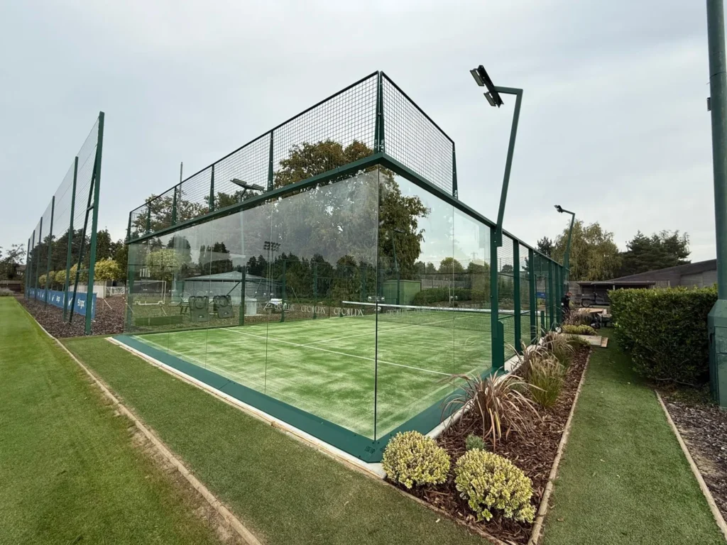 Outdoor padel court with glass walls, green turf, perimeter fencing, and overhead lighting
