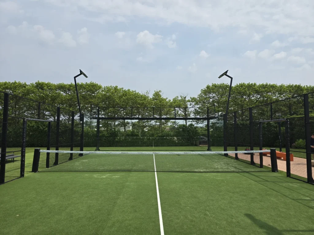 Outdoor padel court with green turf, central net, perimeter fencing, and overhead lighting