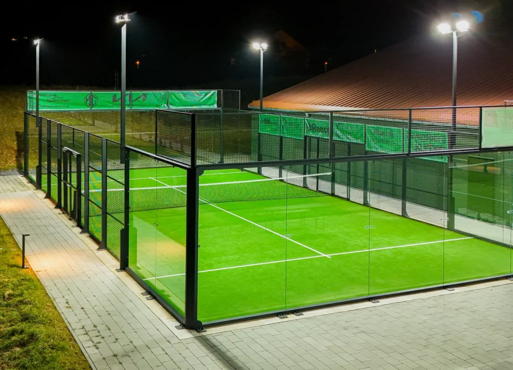 Outdoor padel court with green turf, glass walls, fencing, and overhead lighting at night